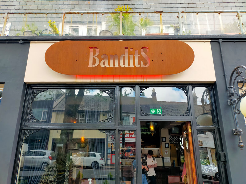 Picture shows the front of Bandits, the cafe sign and a its door and windows, with some artistic and unplanned reflections from the street, including cars and a tree.