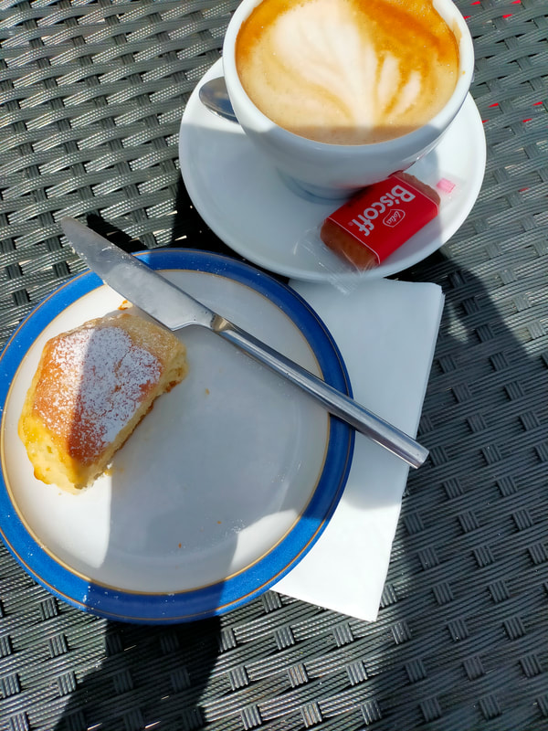 Picture shows a cup of coffee with a saucer, and a half-eaten plain scone. The shadow of the photo-taker falls across the table.